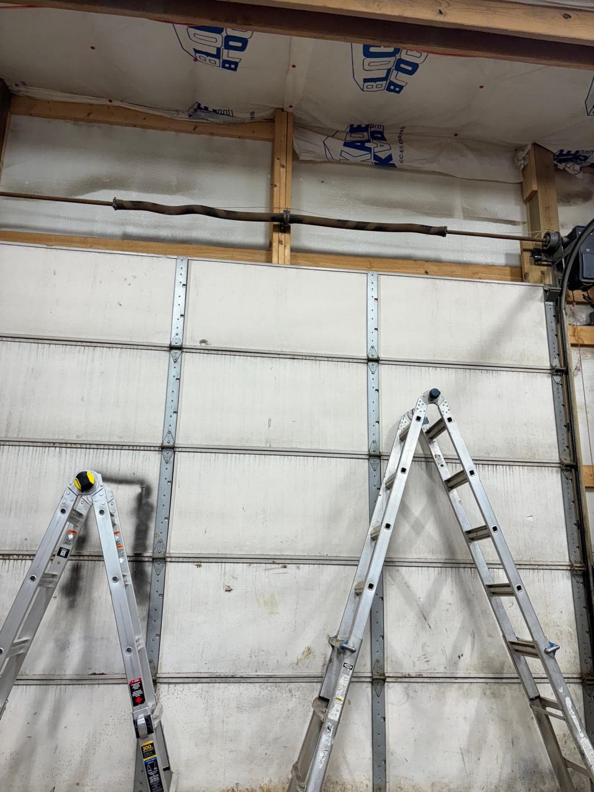 Inside a pole barn during door work: a tall white sectional door with two stepladders set up against it and the metal framing above
