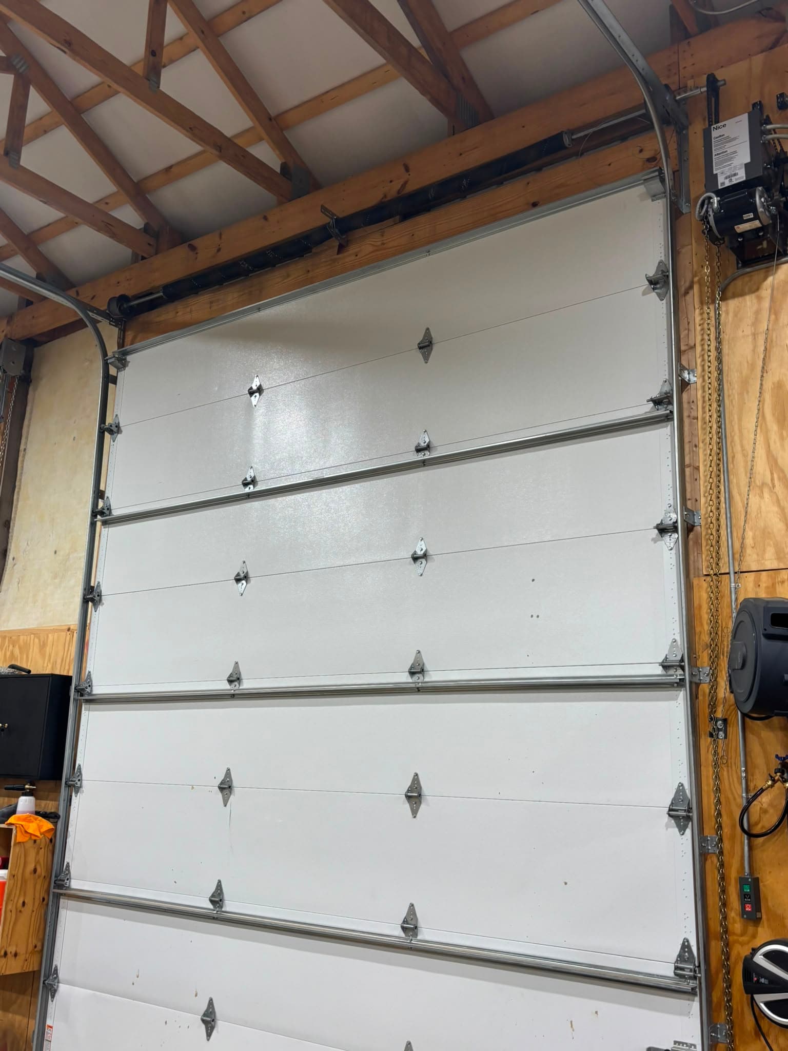 The inside face of a tall white sectional door in a pole-barn style building, showing the hinges, rollers, vertical track and the torsion spring on the shaft above the opening, with exposed timber trusses overhead