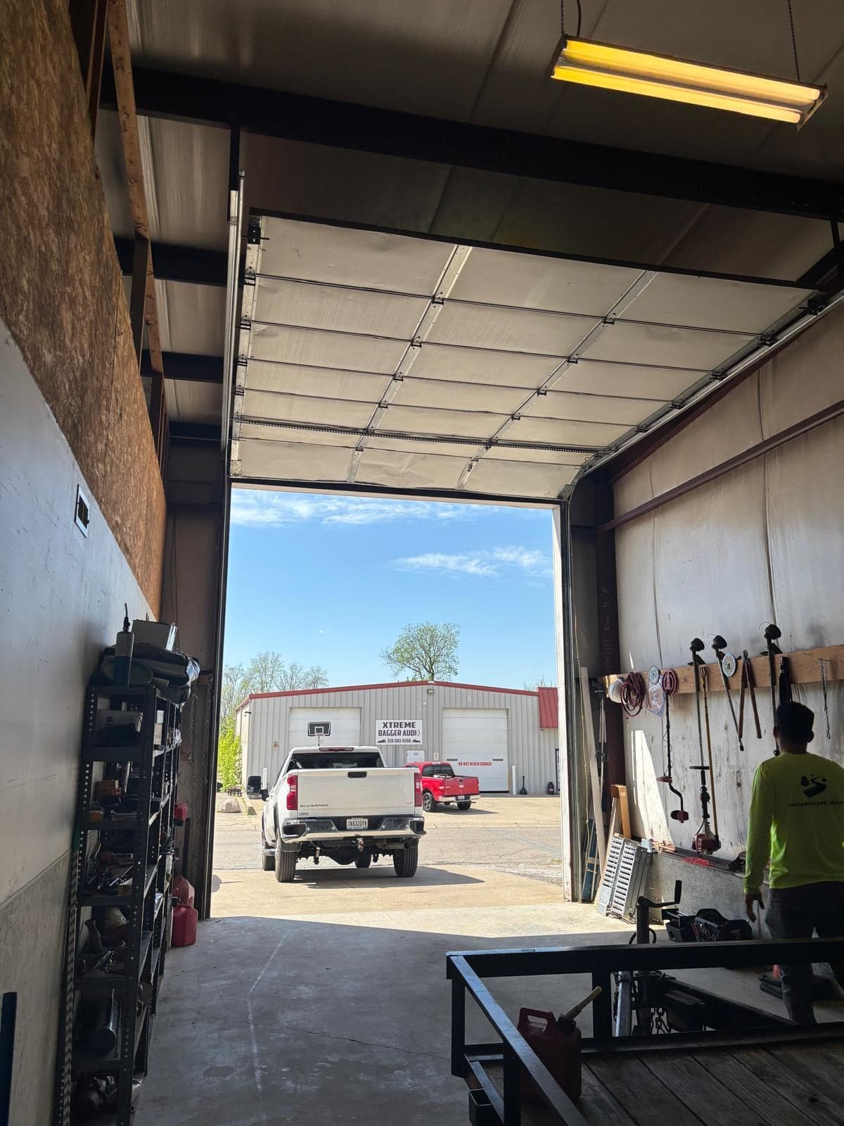 Inside a commercial shop with the bay door open, a pickup truck and another building outside and a technician working at the back