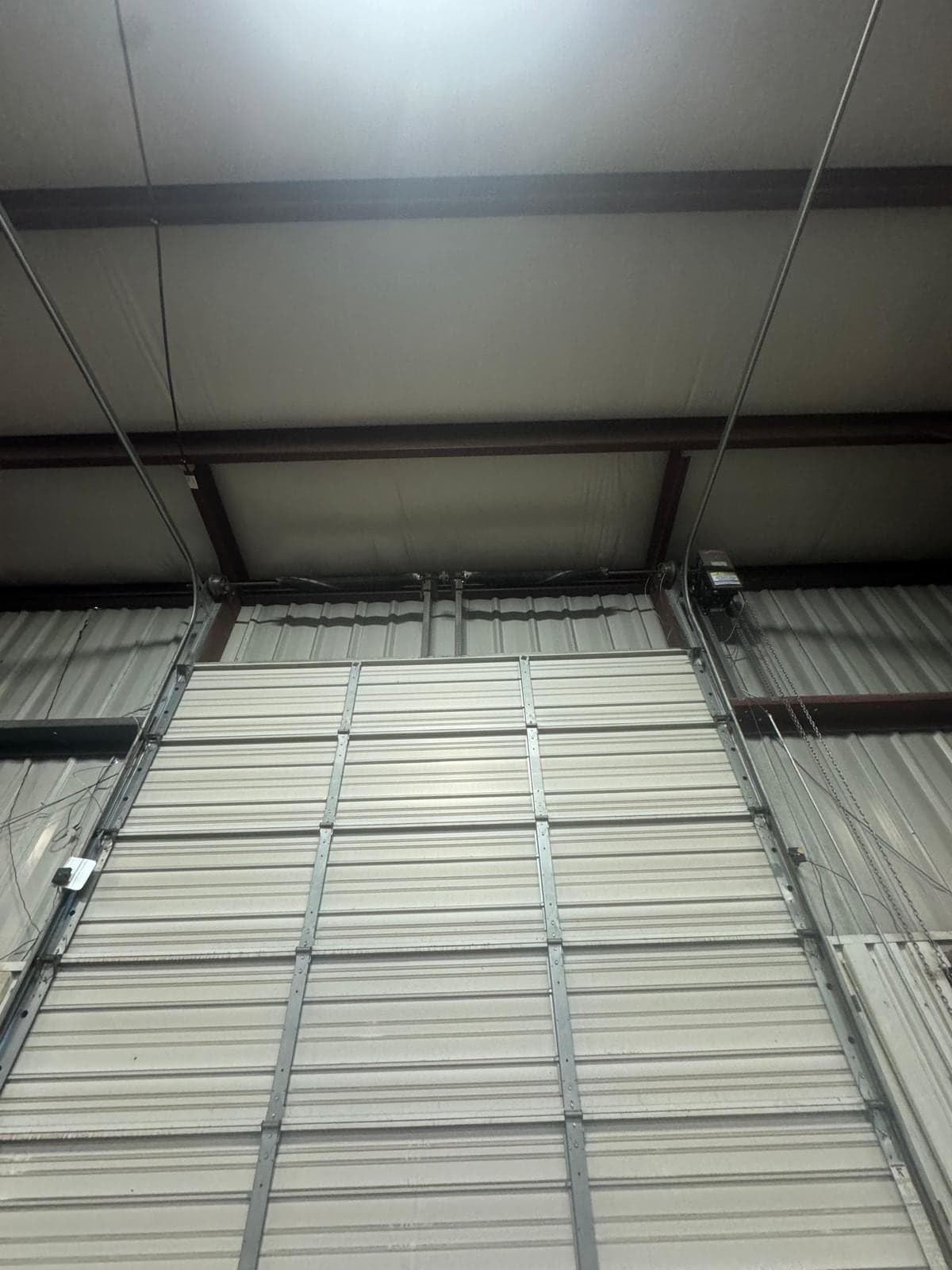 Looking up at a tall white sectional door and the exposed roof structure inside a steel building