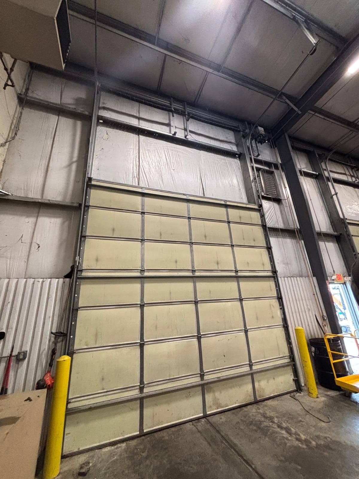 A large white sectional door inside a steel building, with yellow safety bollards at the floor and the roof structure overhead