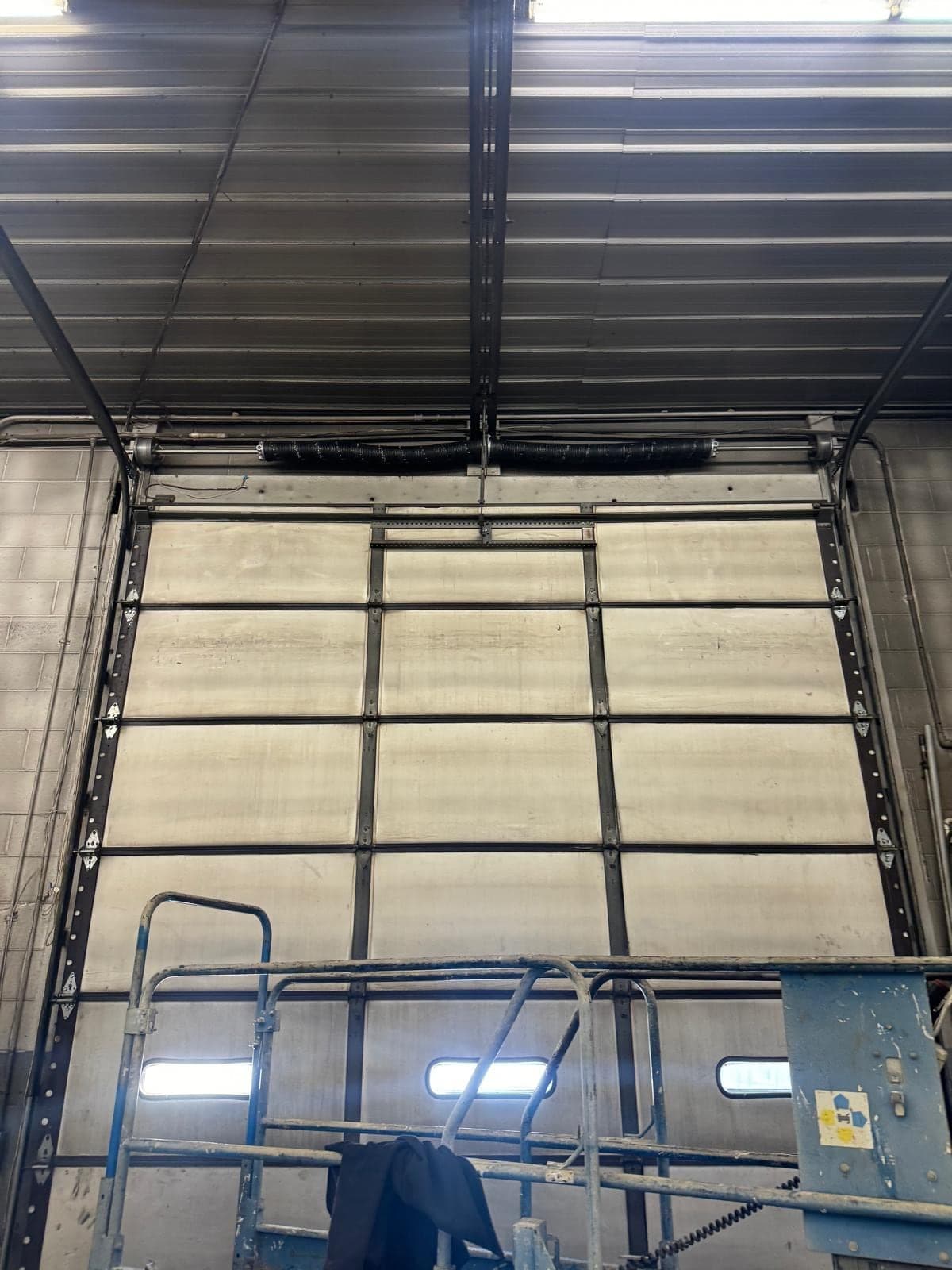 A tall industrial sectional door seen from inside a steel building, with its vertical tracks running high up the wall and a small view out at the bottom