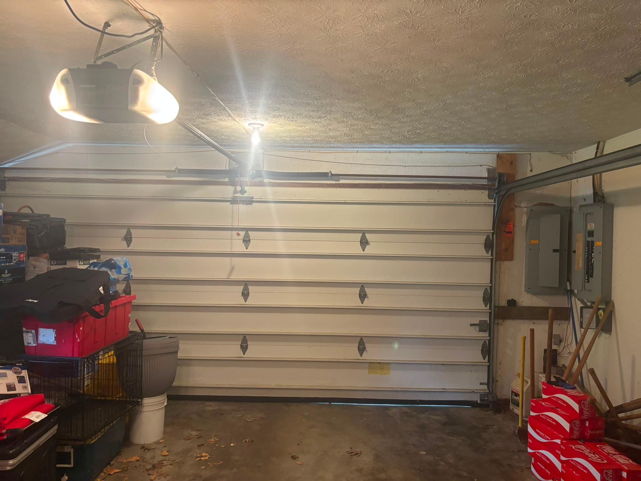 The inside of an attached garage with the door closed, showing the ceiling-mounted opener with its light on, the torsion shaft and spring running above the door opening, and the household's stored belongings along the wall