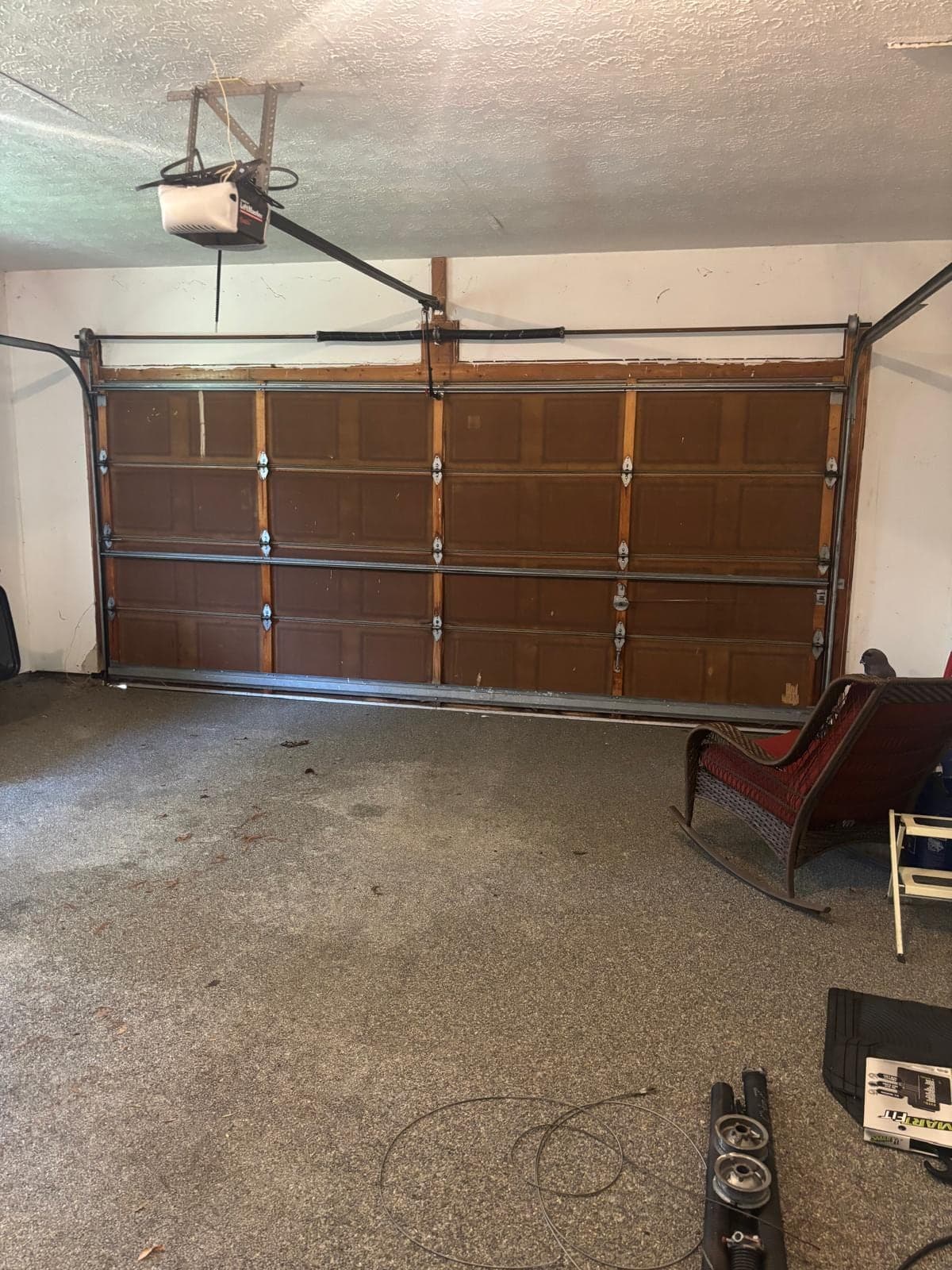 A brown raised-panel garage door seen from inside a single-car garage, with the torsion shaft and opener rail above it and a rocking chair stored against the wall