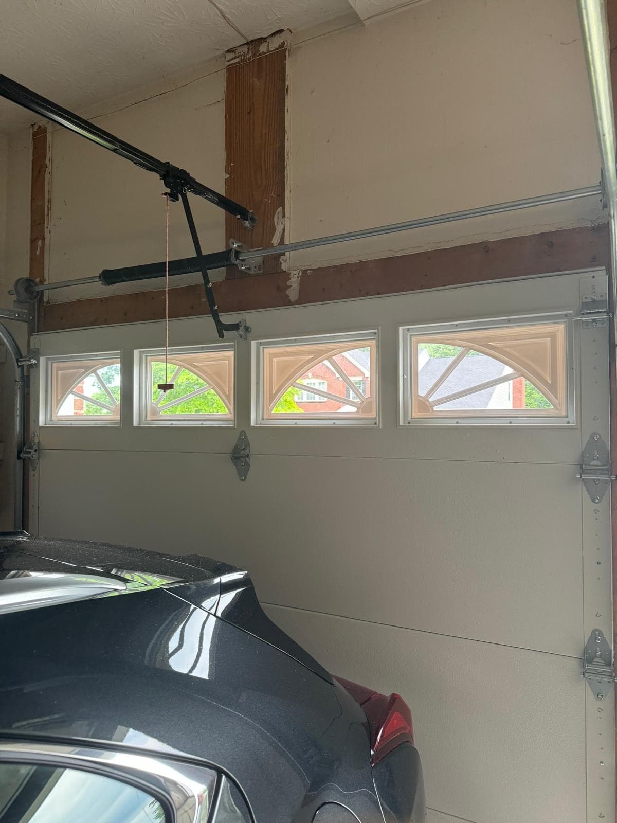 Inside an attached garage: a white sectional door with arched windows, the opener rail and emergency release cord overhead, and the hood of a car in the foreground