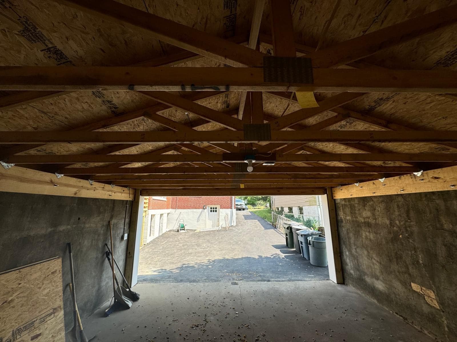 An older detached garage seen from inside with the door open, exposed roof trusses and ceiling joists overhead and the driveway and trash bins visible outside