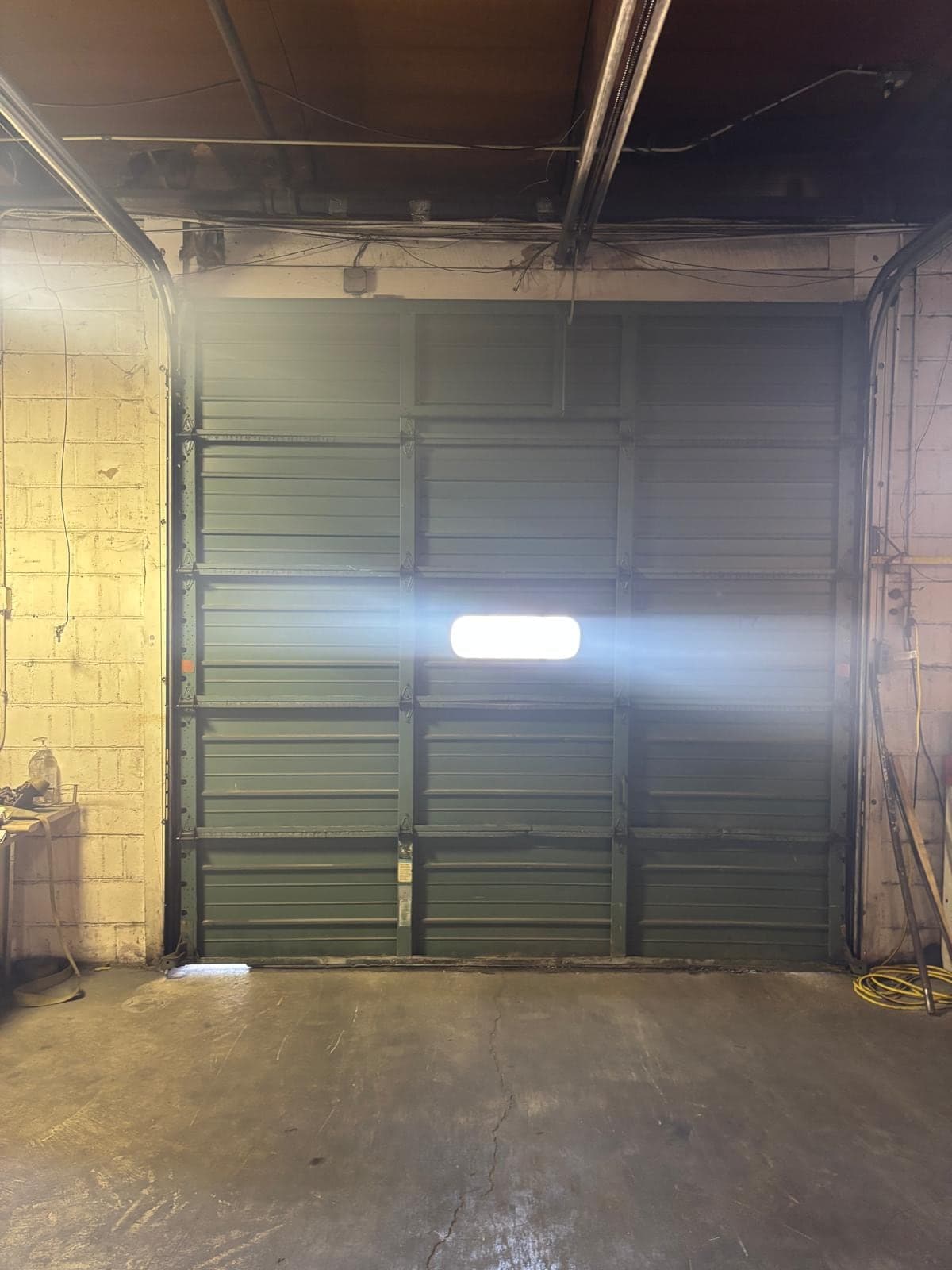 A dark older steel garage door inside a concrete-block garage, lit by a single bright work light in the centre of the door