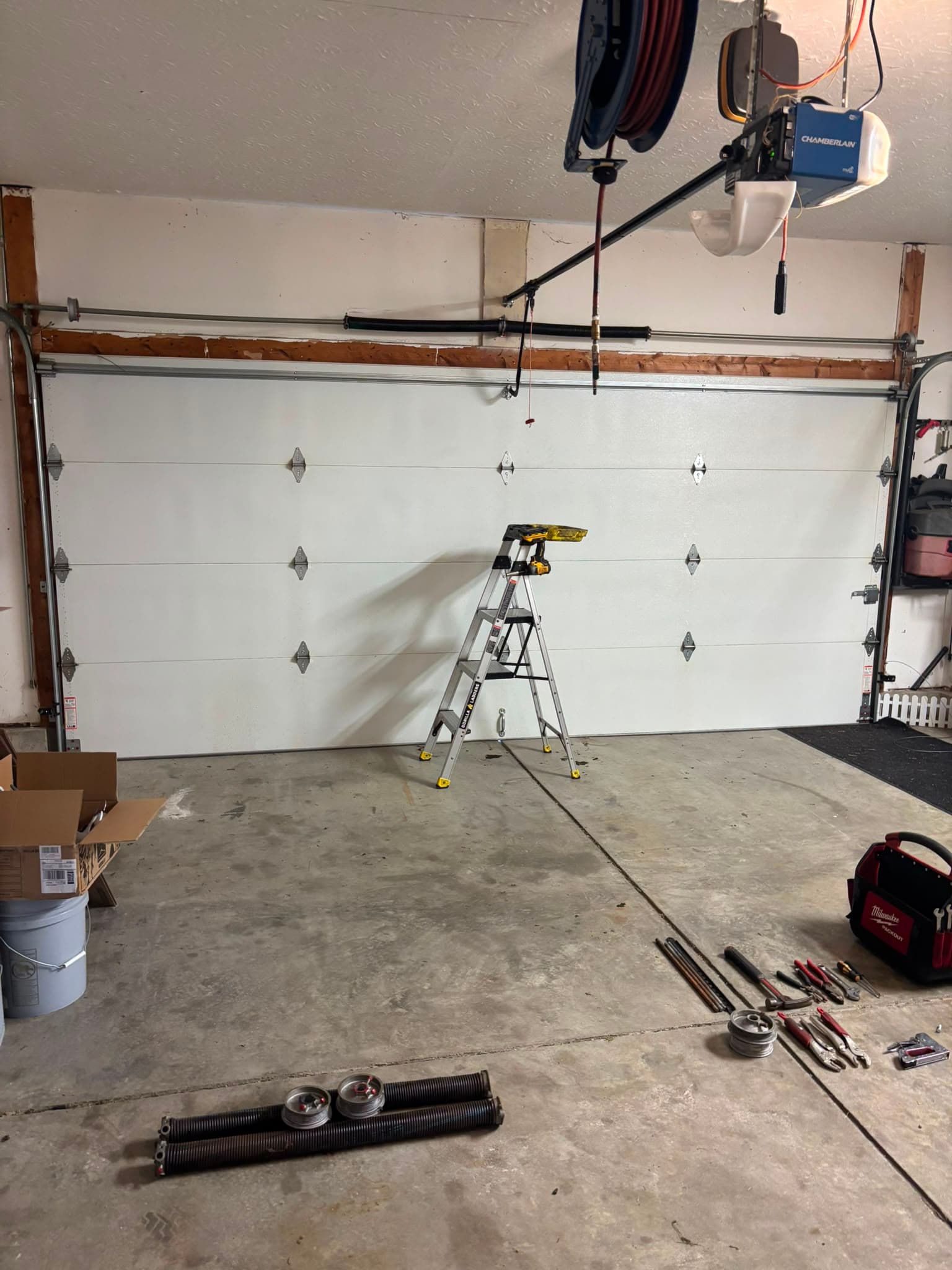 A torsion spring replacement part-way through: two removed springs and their cable drums laid out on the garage floor alongside winding bars and hand tools, with a Chamberlain opener overhead