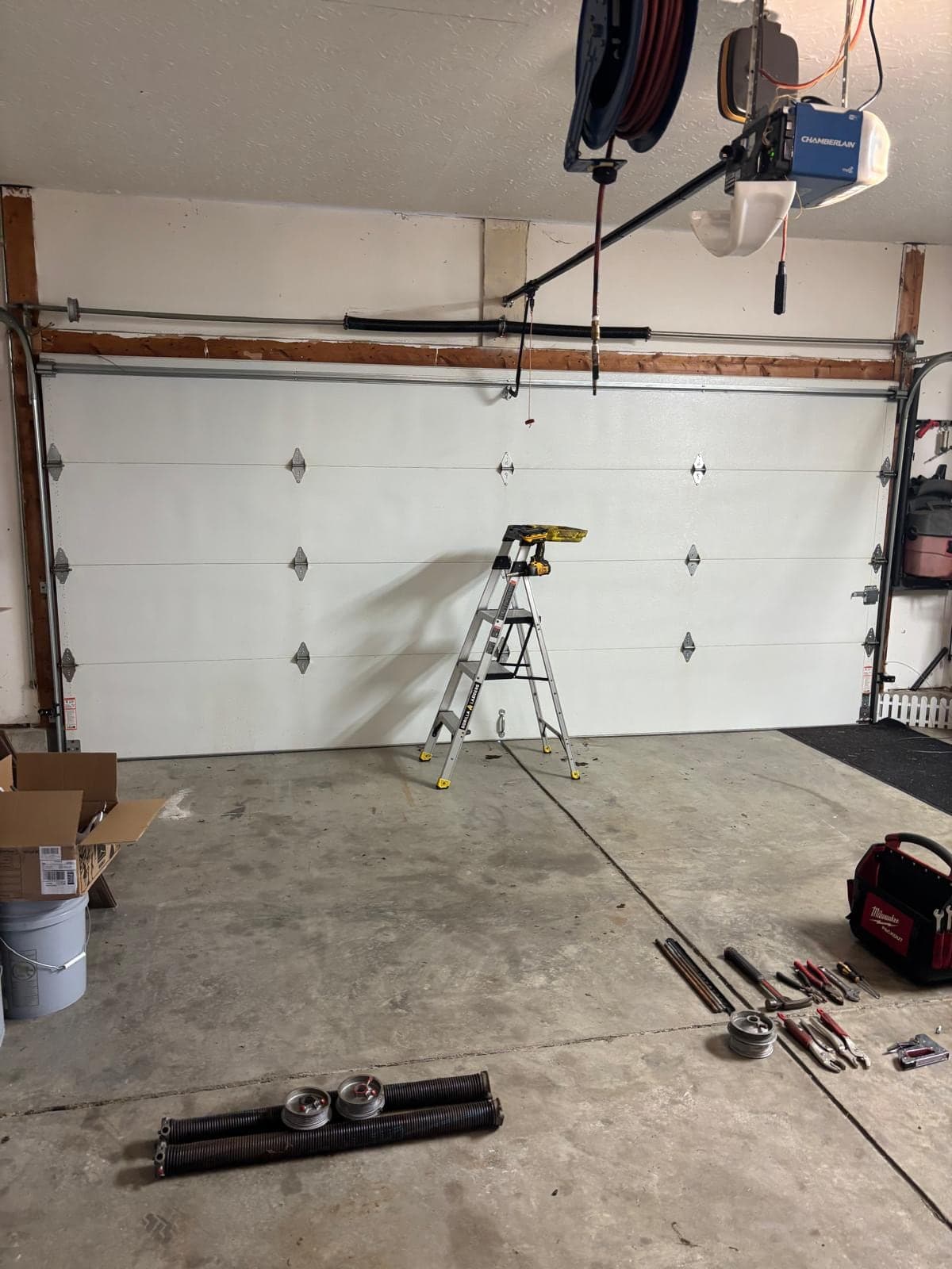 Inside a garage during installation: a new white sectional door in the opening, a stepladder in the centre, torsion hardware going up and tools laid out on the floor