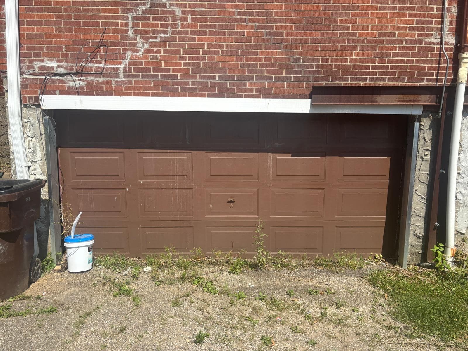 Two old brown raised-panel garage doors in a brick building, with overgrown grass and a white bucket at the base
