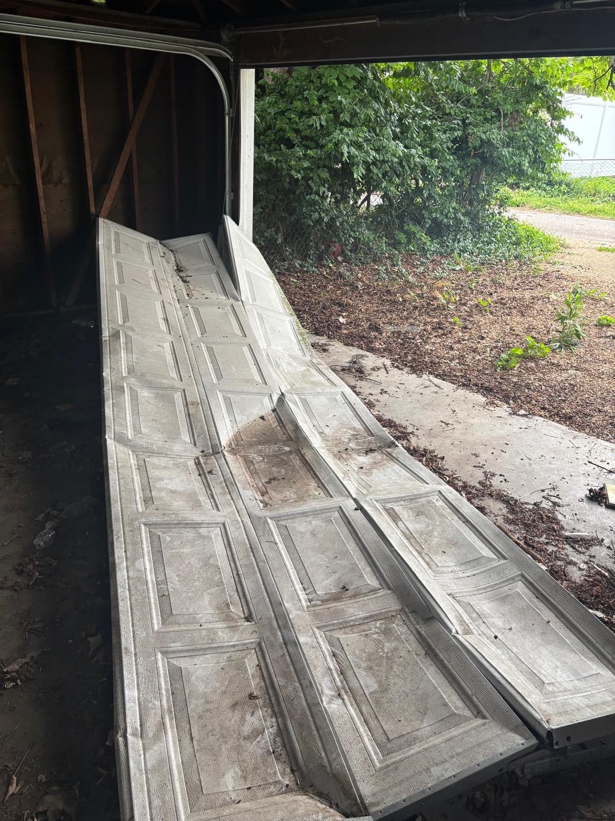 Old white garage door sections lying outside an open garage after removal, with the inside framing and the yard beyond