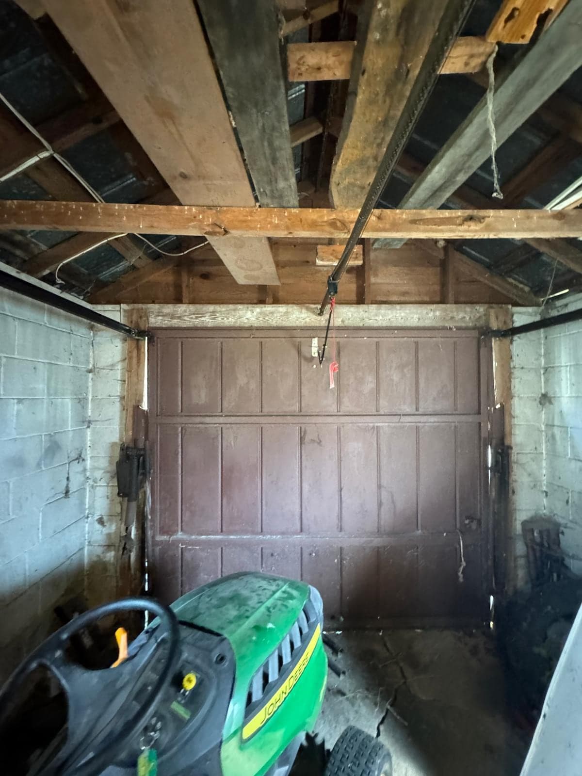 An old dark-brown wood garage door inside a concrete-block garage with exposed roof timbers and a green riding mower parked in front