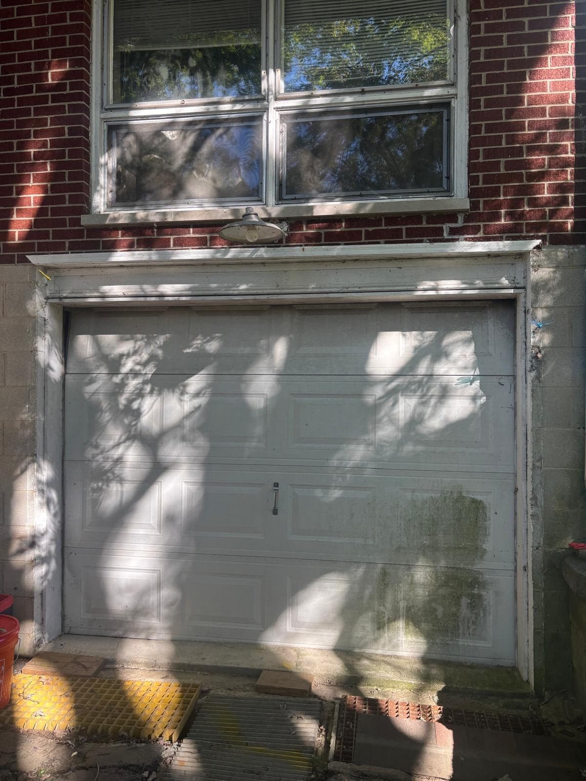 An older white single garage door with dirt streaks and a scuffed bottom edge, set in a brick wall below a window