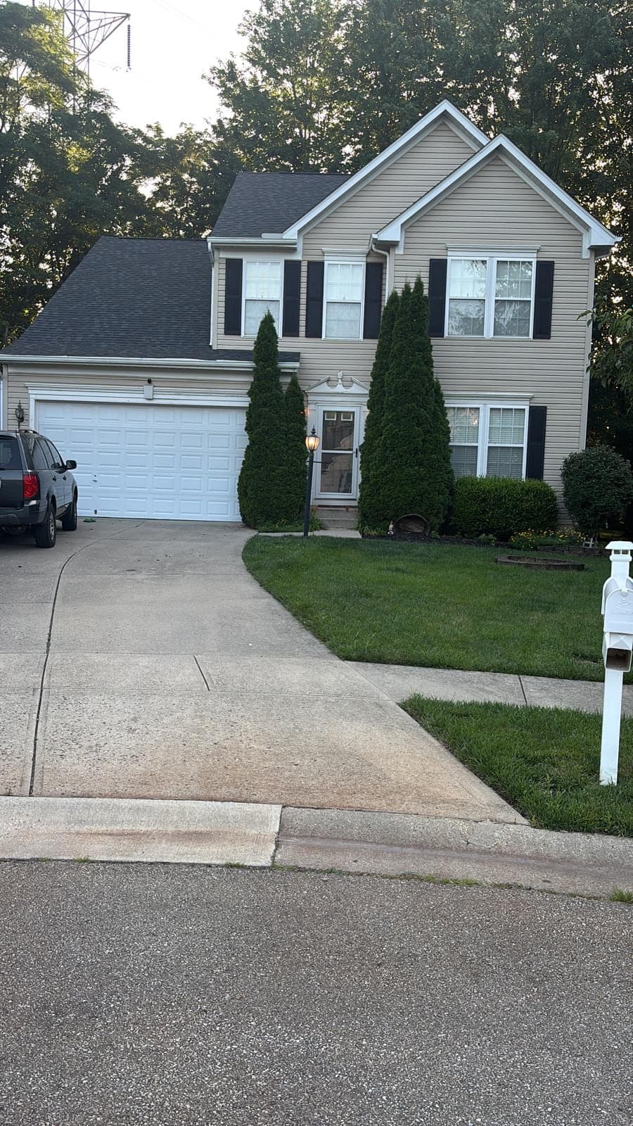 A two-storey house with grey vinyl siding and brick lower walls, a white double garage door, trimmed evergreens by the entrance and a long concrete driveway