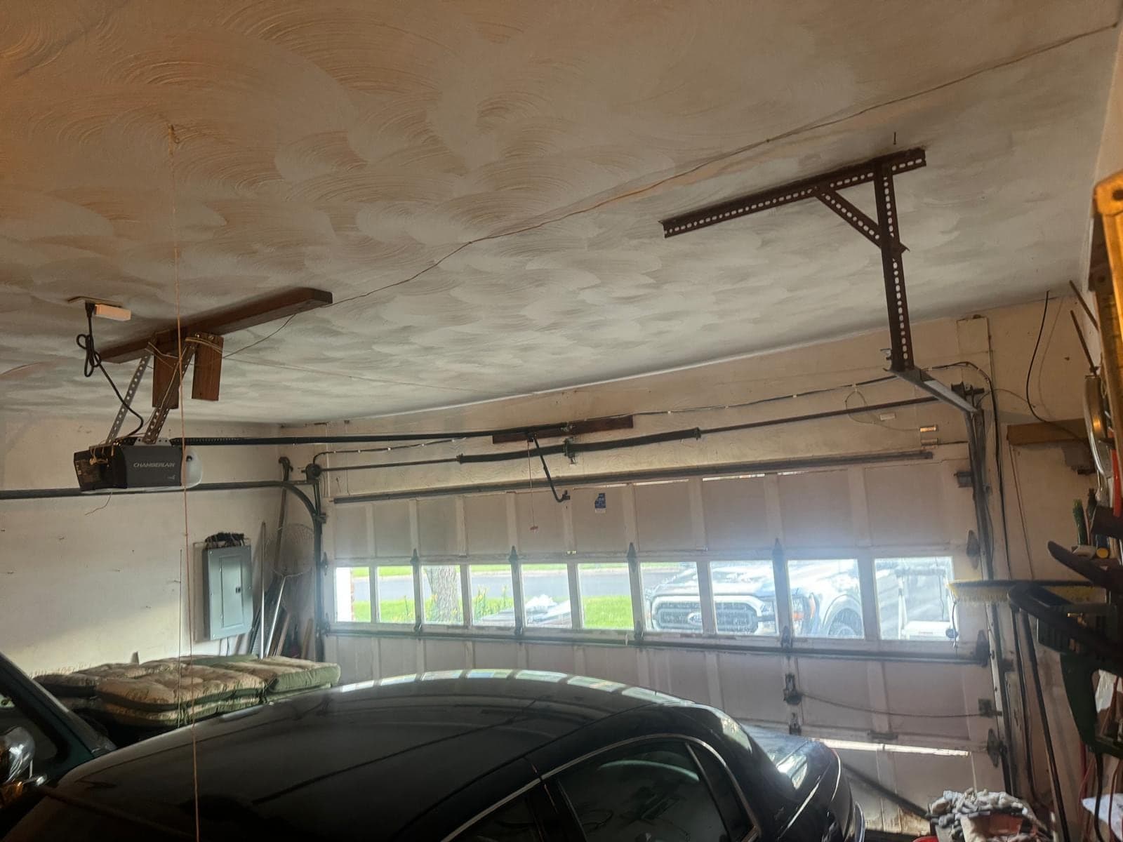 A garage ceiling with a mounted opener and rail, the top of a white door with a row of windows, and a dark car parked underneath