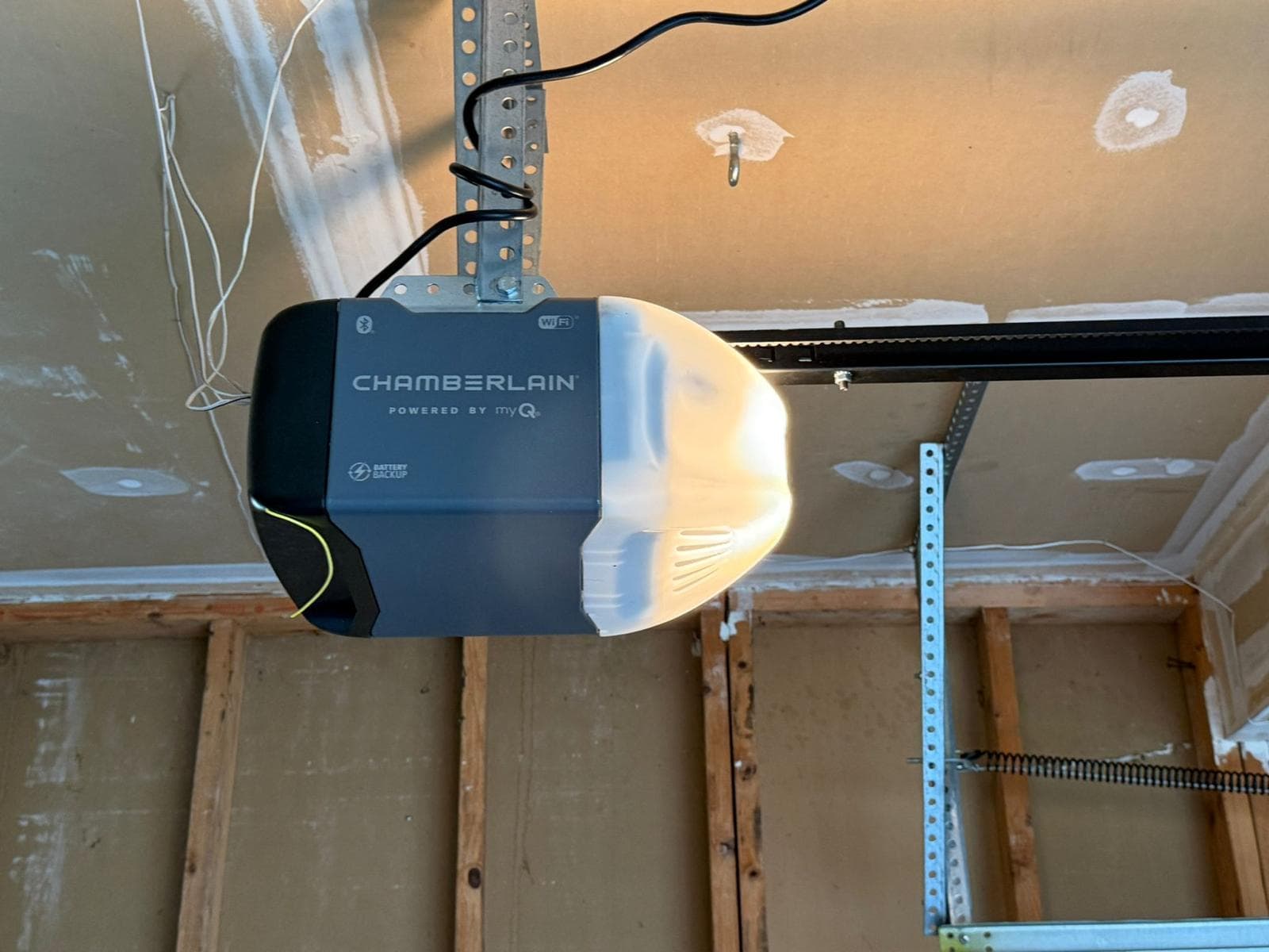 A newly installed Chamberlain myQ garage door opener mounted to ceiling framing, with its light on, the belt-drive rail running off to the right, and Wi-Fi, Bluetooth and battery-backup badges visible on the black housing