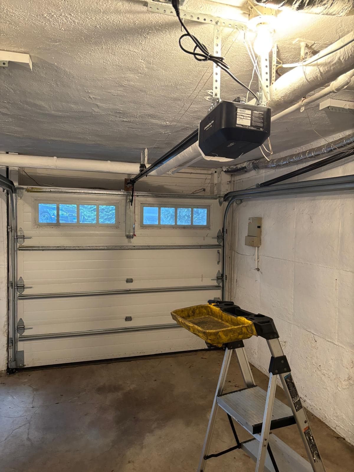 Inside a garage with a white sectional door that has glass-block style windows, a new ceiling opener and rail overhead and a step stool with tools on the floor