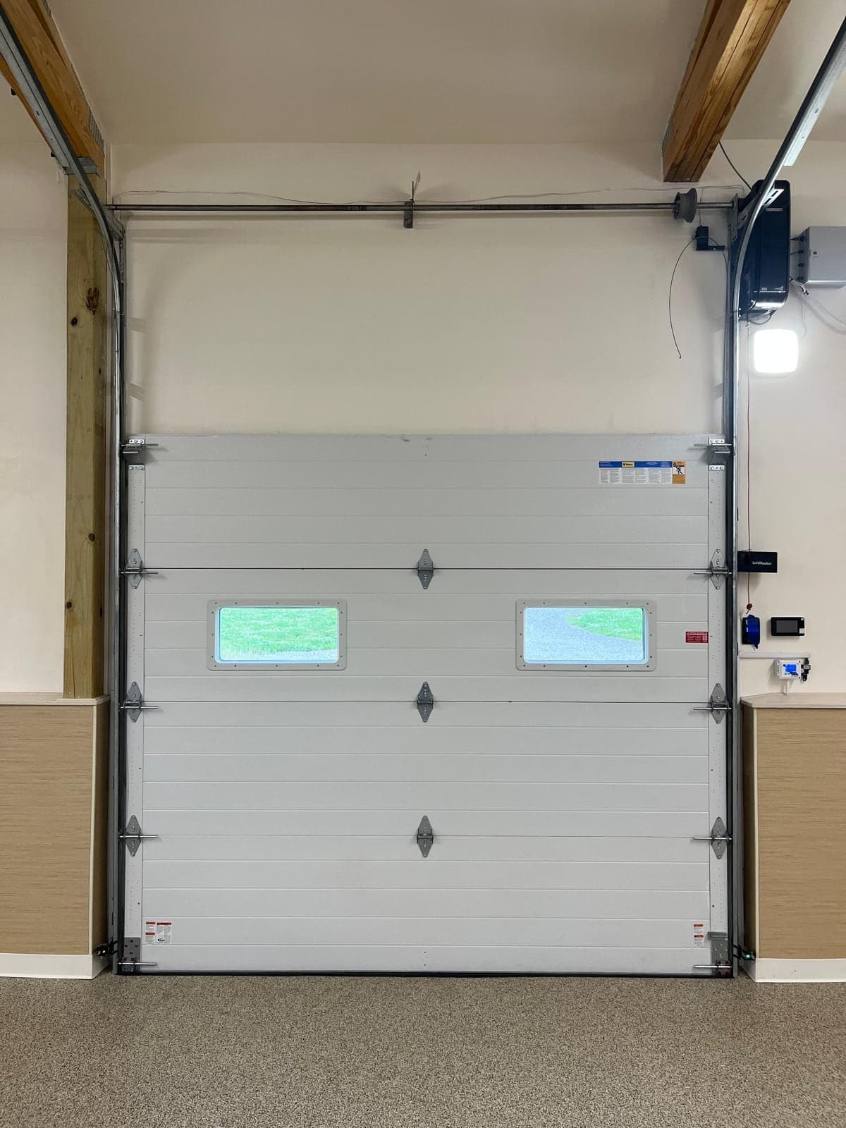 A freshly installed white sectional garage door with two small windows, a new ceiling opener with its light on and the wall control beside the door