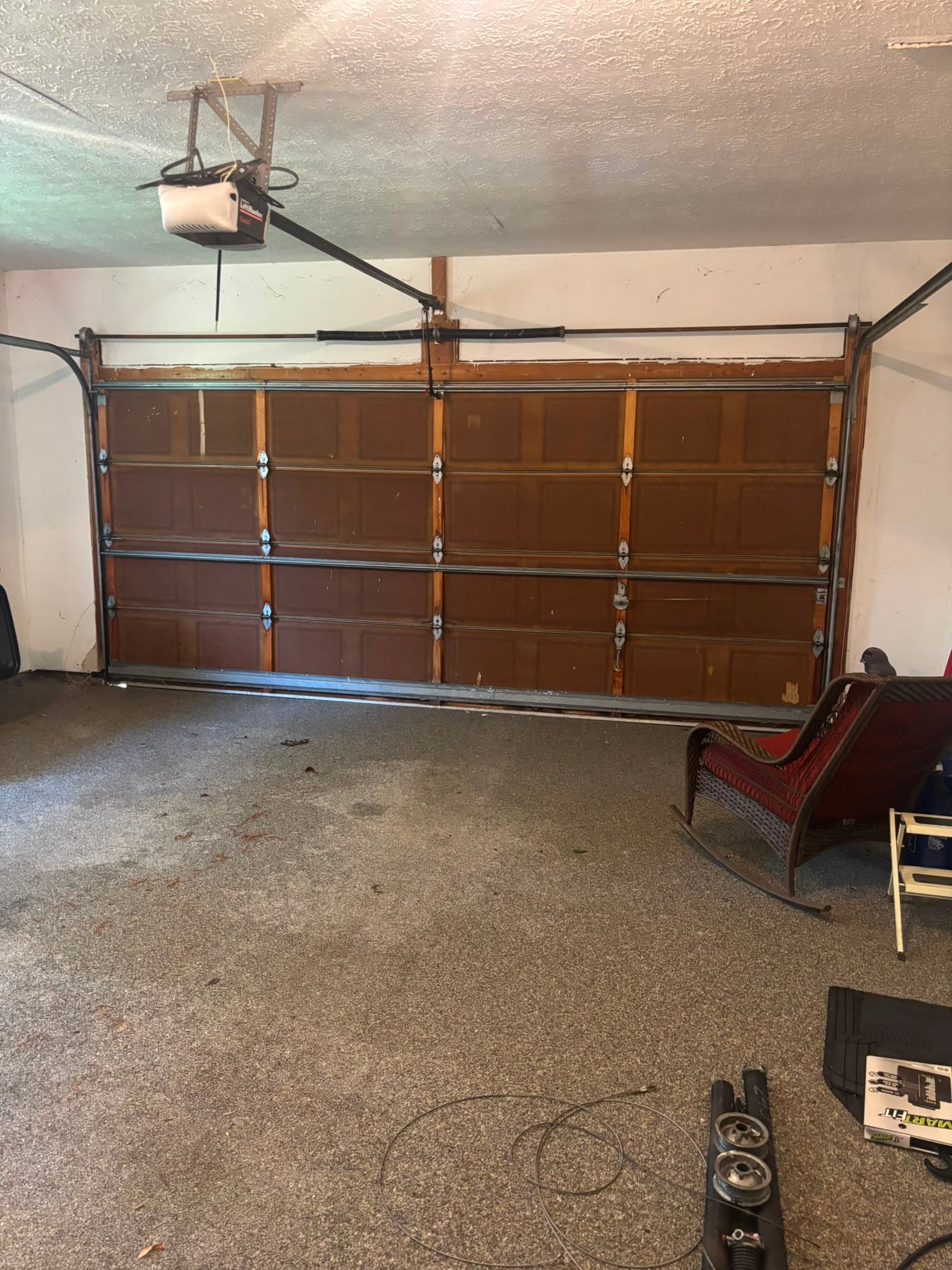 An older white LiftMaster chain-drive opener mounted on a ceiling bracket above a brown wood raised-panel garage door, with the torsion shaft across the opening and removed springs, cable drums and a loose lift cable lying on the garage floor