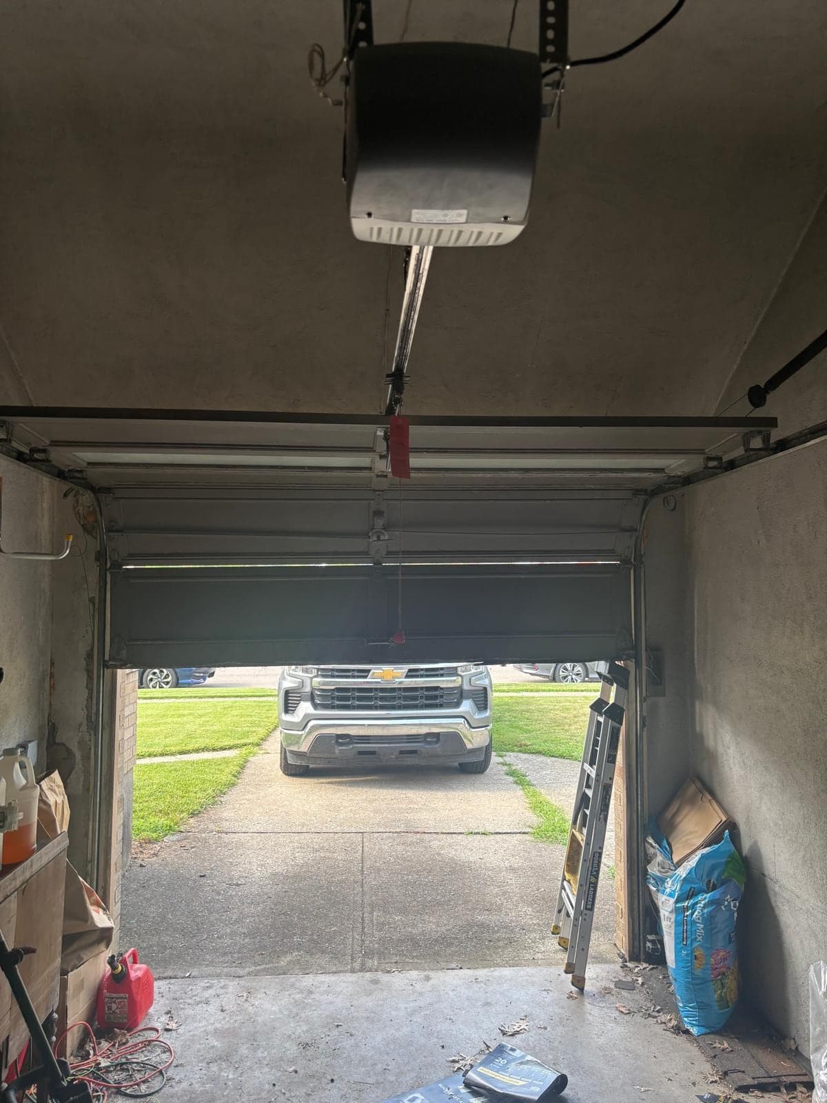 A ceiling-mounted garage door opener and rail seen from inside a small garage, looking out through the open door at a pickup truck in the driveway