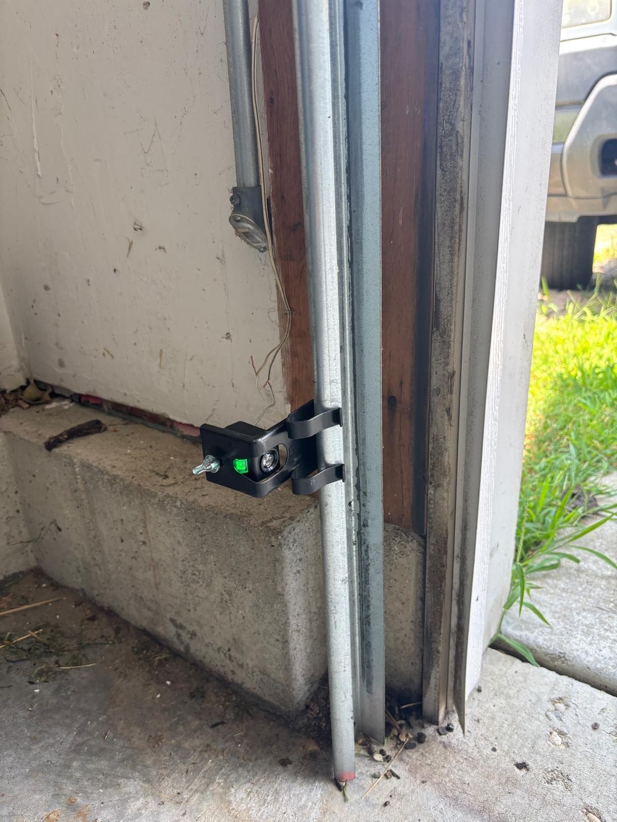 A photo-eye safety sensor on its bracket at the base of a garage door track, with the wiring run down the jamb and grass outside the open door