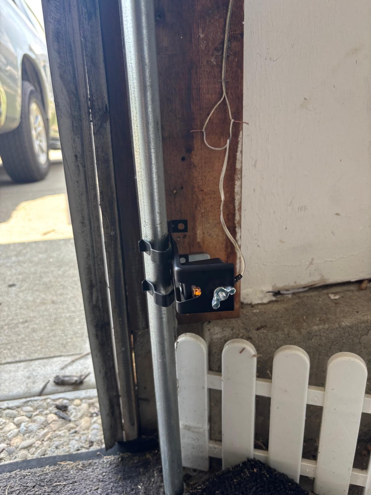 The second photo-eye sensor mounted on the opposite track at floor level, beside a white picket fence outside the open garage