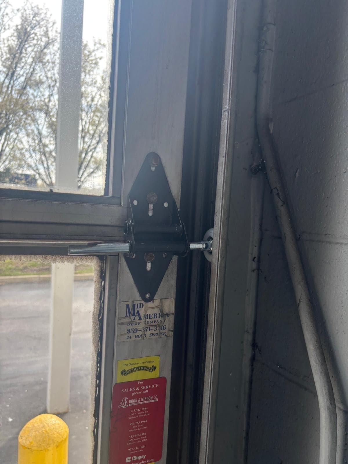 Close-up of a steel hinge and roller bracket on the edge of a sectional door, with a supplier sticker on the track and a driveway outside