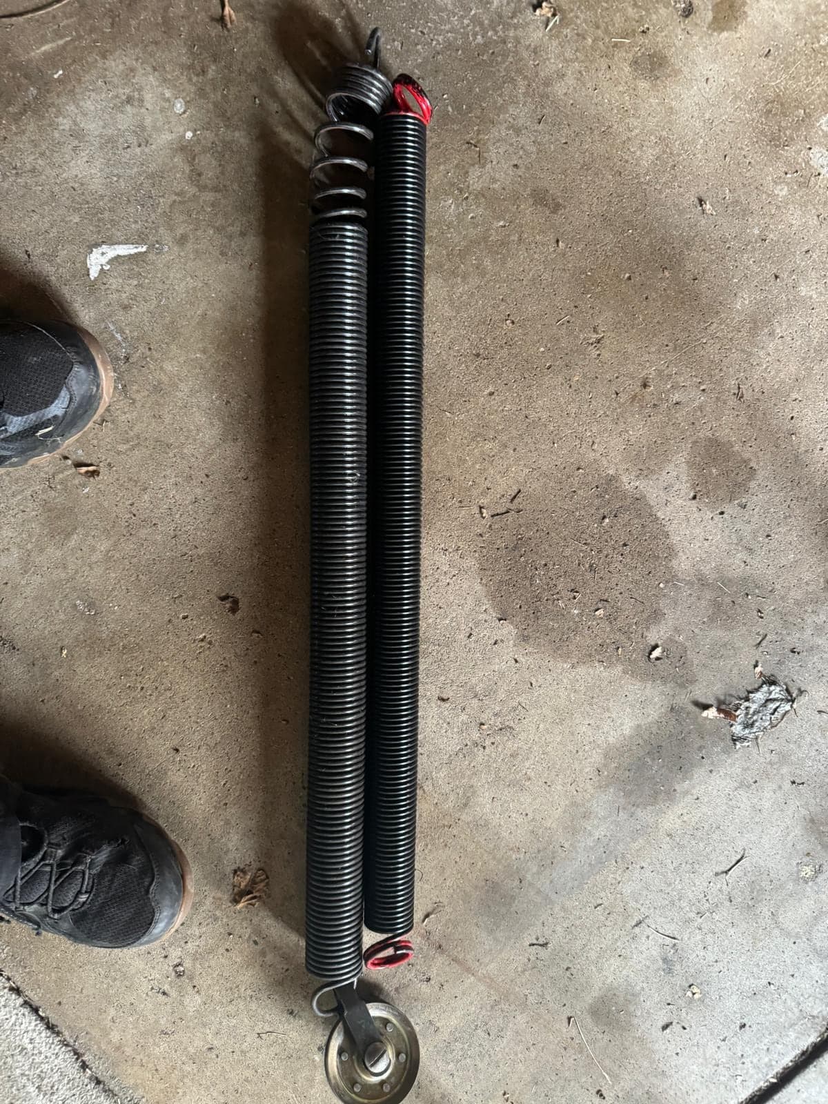 A new black torsion spring lying on a concrete garage floor between a technician's boots, still in its shipping wrap