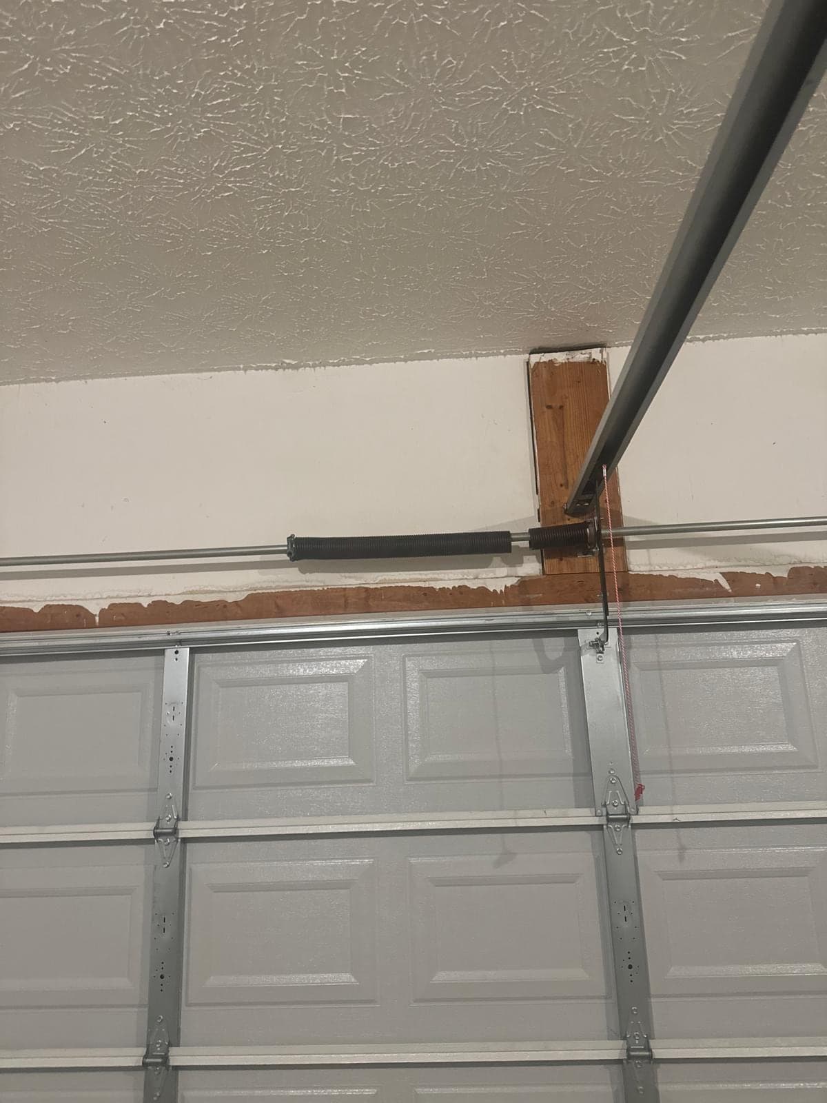 The top section of a grey raised-panel door seen from inside, with the torsion shaft, centre bearing bracket and horizontal track where they meet the ceiling